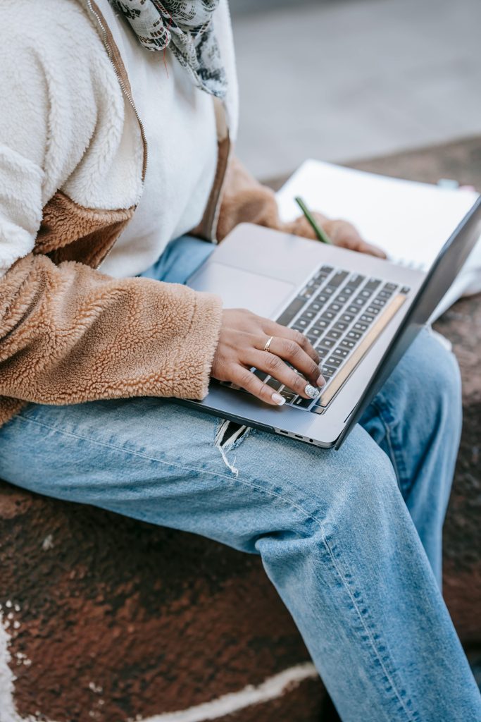 From above of crop unrecognizable lady in stylish clothes typing on laptop keyboard while sitting on bench in campus and preparing for exams